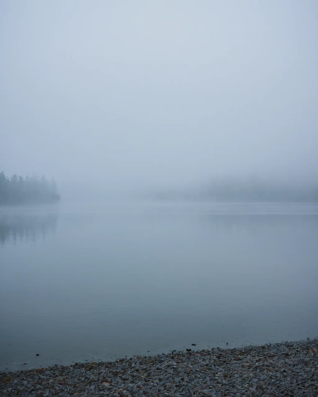 A foggy lake with a rocky shore and distant trees barely visible through the mist.
