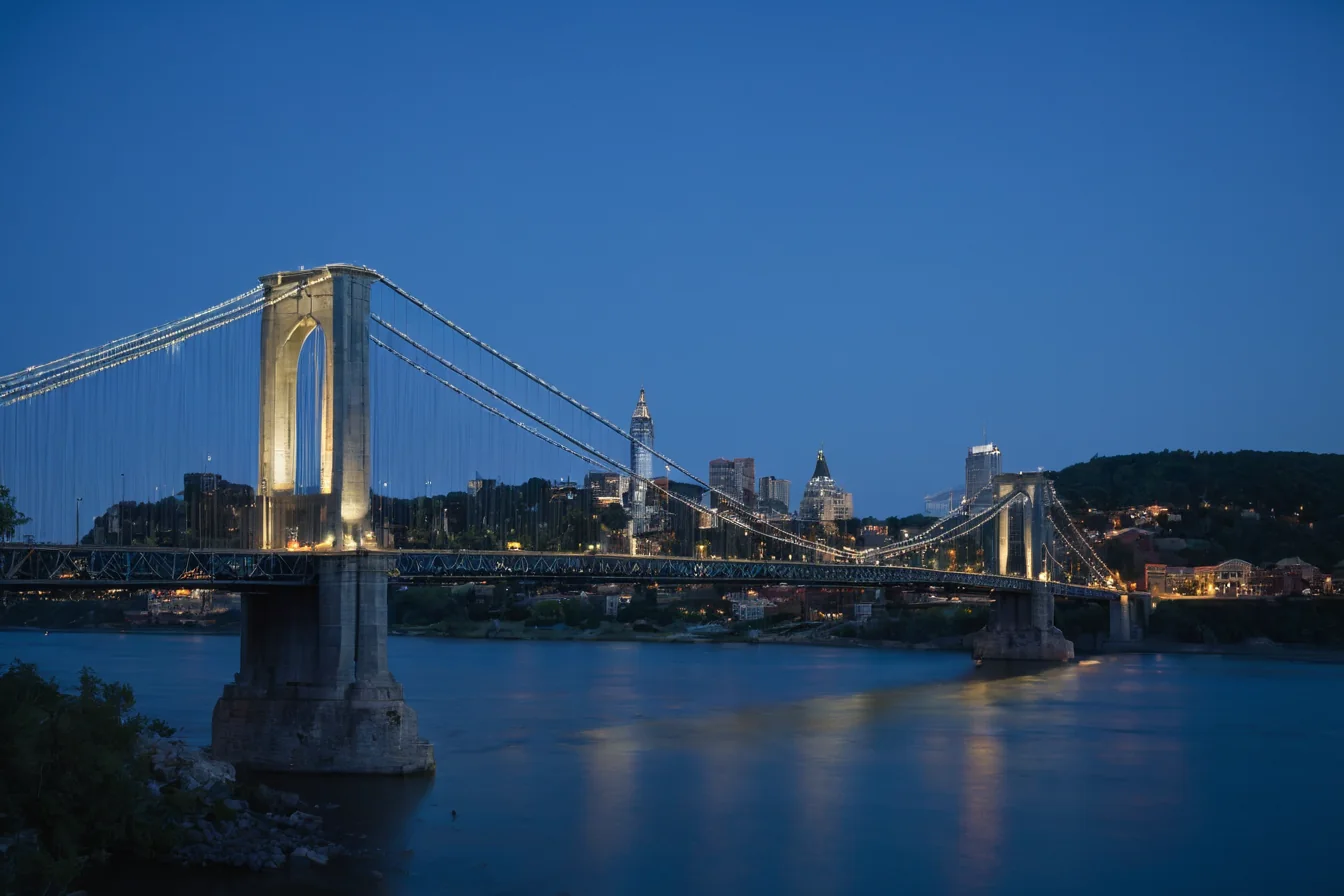 A lit suspension bridge spans a river with a city skyline in the background at dusk.