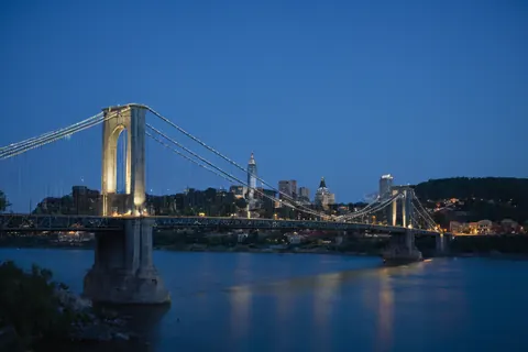 A lit suspension bridge spans a river with a city skyline in the background at dusk.