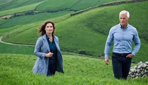 A man and woman walking on a grassy hill with stone walls and winding paths.