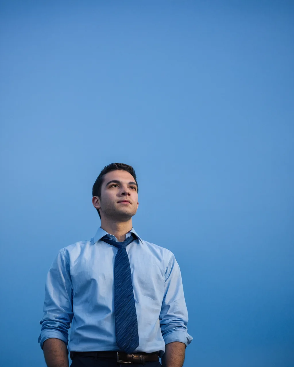A man in a blue shirt and tie looking up against a solid blue background.