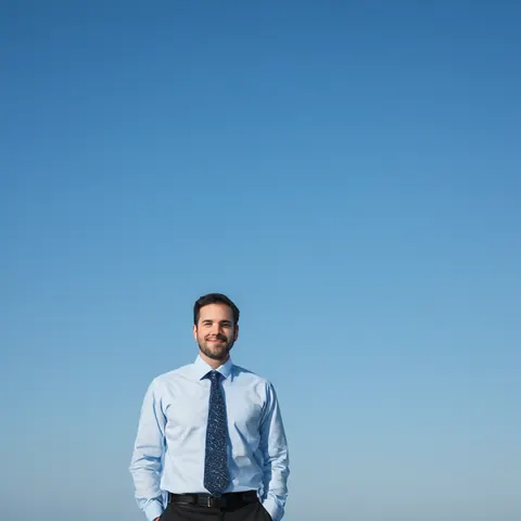 A man in a blue shirt and tie smiling against a clear blue sky