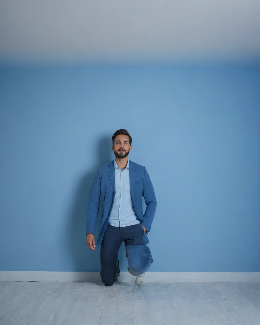 A man in a blue suit kneeling against a blue wall with white floor.