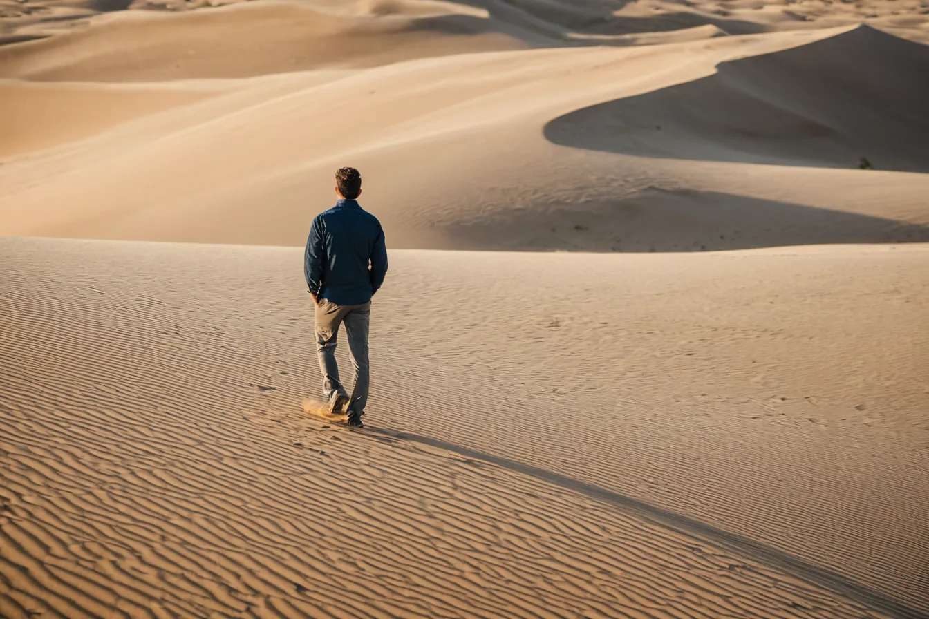 A man walking alone across textured sand dunes in a desert at sunset