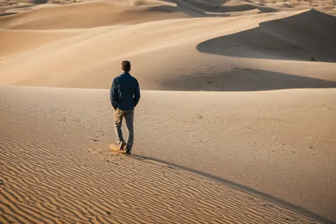 A man walking alone across textured sand dunes in a desert at sunset