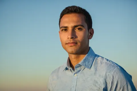 A man wearing a light blue shirt standing outdoors against a clear sky during golden hour.