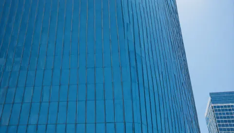 A tall blue glass building curves upward against a clear sky with another building nearby.