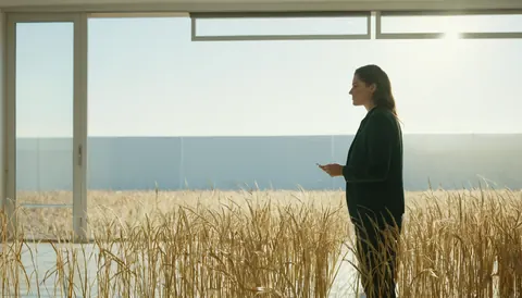 A woman in a dark suit stands inside a glass-walled structure surrounded by tall golden wheat stalks under bright sunlight.