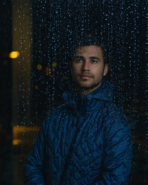Man in blue jacket looking through rain-streaked window at night with blurred lights behind him.