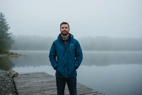 Man in blue jacket standing on wooden dock beside misty lake with trees in background