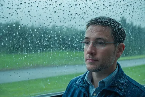 Man in glasses looking out rain-streaked window at green landscape