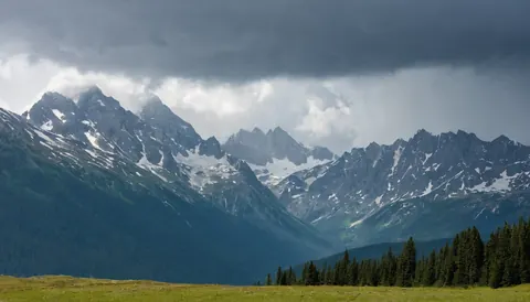 Snow capped mountains under dark storm clouds with green meadow and pine trees below