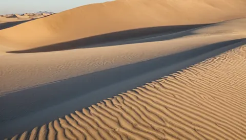 Sunlit desert dunes with rippled sand and long shadows from distant mountains.