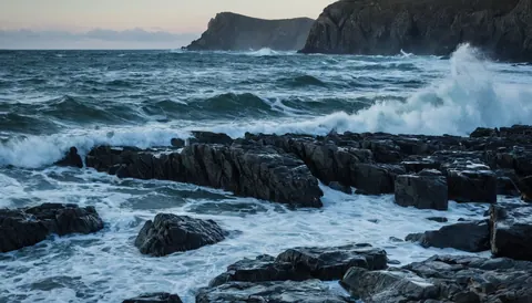 Waves crash against dark rocks at a stormy coastal cliff during dusk