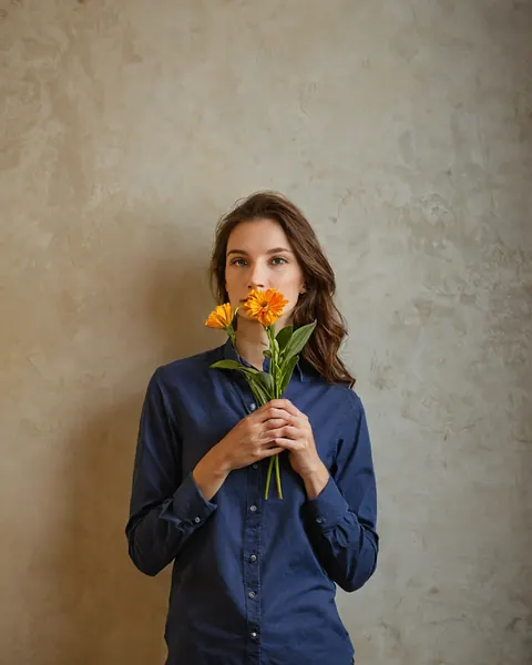 Woman in navy shirt holding orange marigolds to lips against textured beige wall